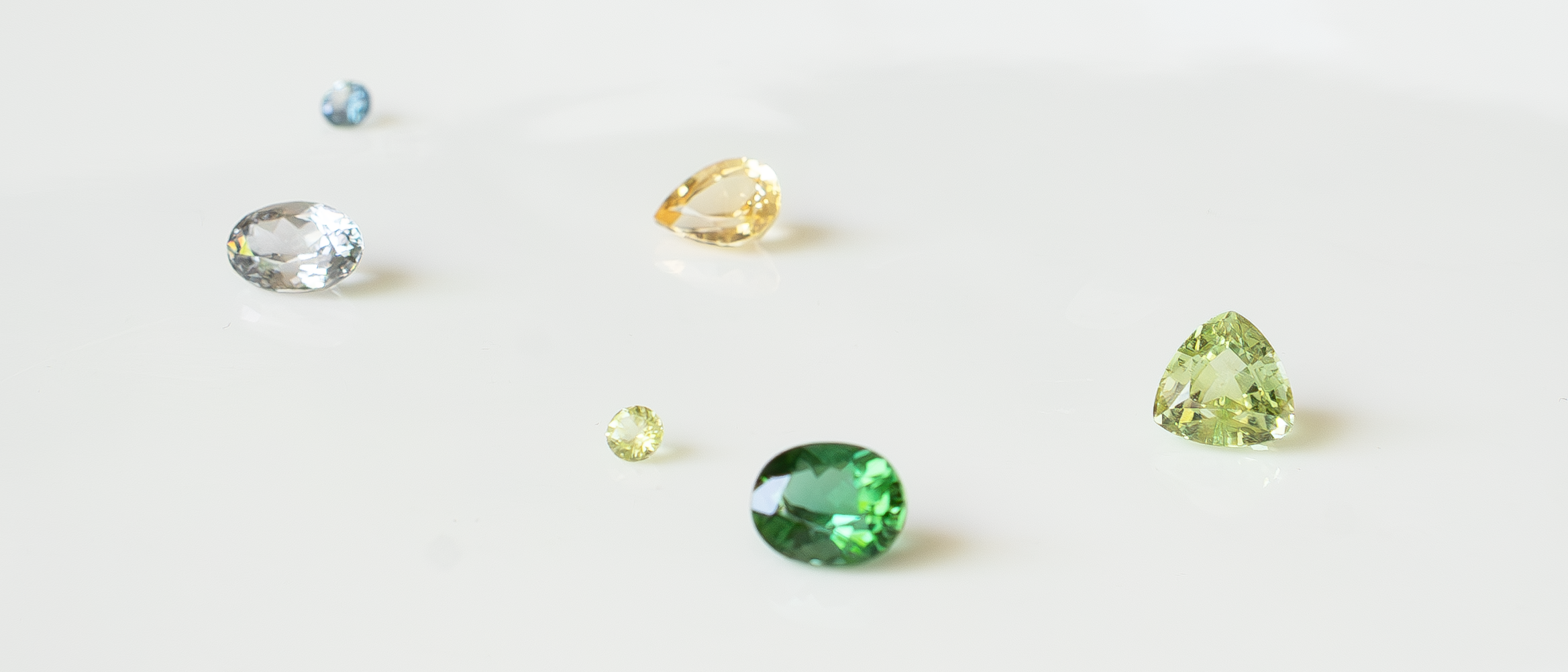 A collection of loose natural gemstones arranged on a soft white surface, including gray spinel, green tourmaline, golden topaz, a small blue sapphire, and a small peridot in various cuts and sizes.