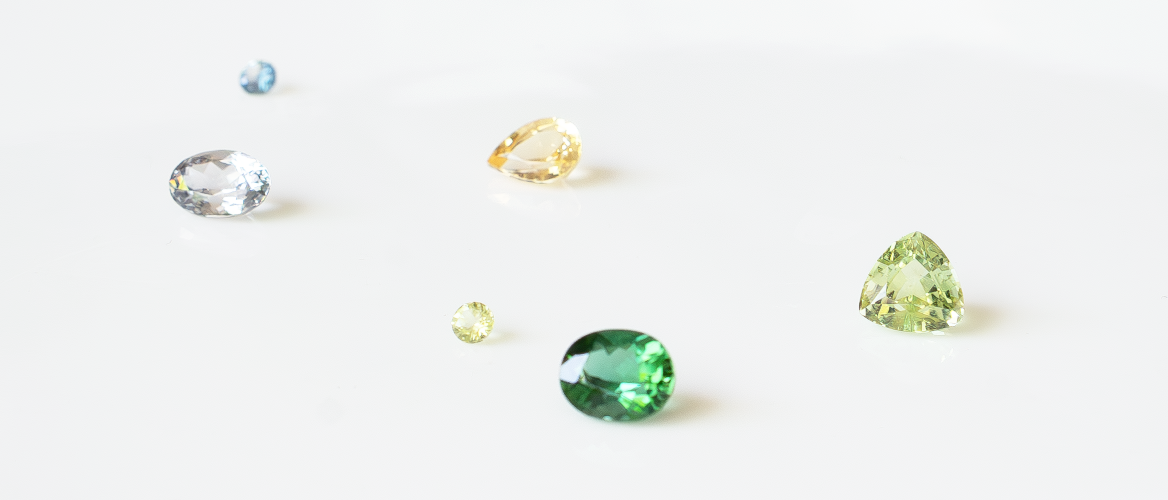 Loose faceted tourmaline, topaz, sapphire, peridot, and spinel gemstones in green, yellow, blue, and clear tones on a bright white surface.