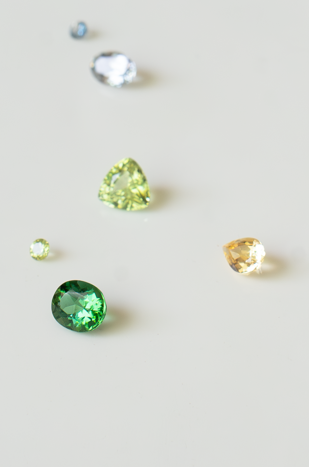 A collection of loose natural gemstones arranged on a soft white surface, including gray spinel, green tourmaline, golden topaz, a small blue sapphire, and a small peridot in various cuts and sizes.