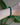 A person's wrist wearing a polished malachite bracelet featuring natural dark green banded stones, 14k gold faceted spacers, and a central diamond-studded gold rondelle. Styled in natural light with tropical greenery in the background.