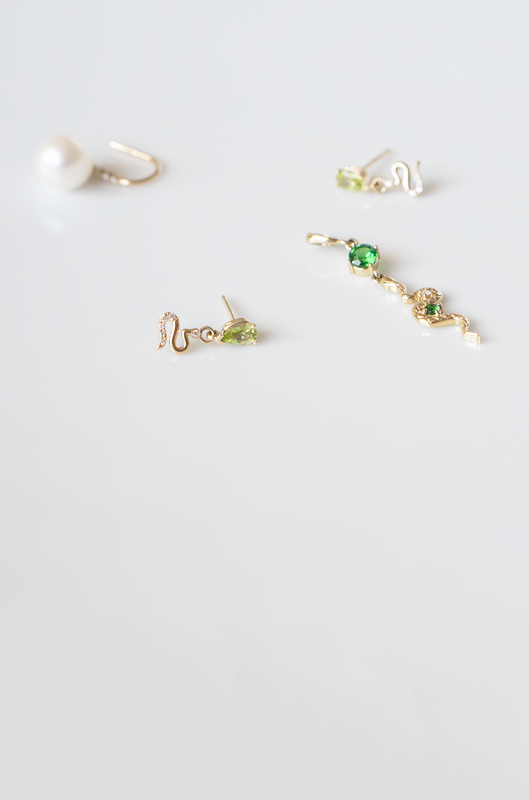 Assorted 14k gold earrings with tsavorite garnet, peridot, diamonds, freshwater pearls, and serpent details on a white reflective surface.