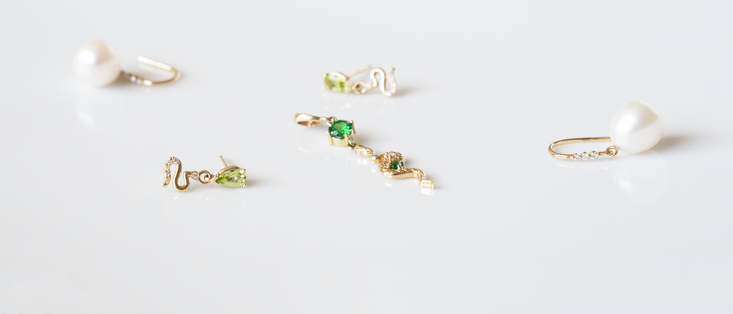 Assorted 14k gold earrings with tsavorite garnet, peridot, diamonds, freshwater pearls, and serpent details on a white reflective surface.